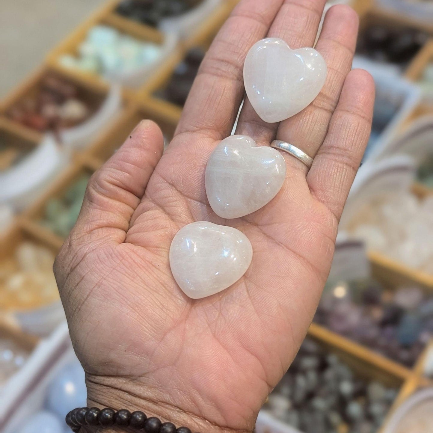 Three polished white heart-shaped crystal pendants are held in a person's open palm, with a blurred shop background showing trays of stones.