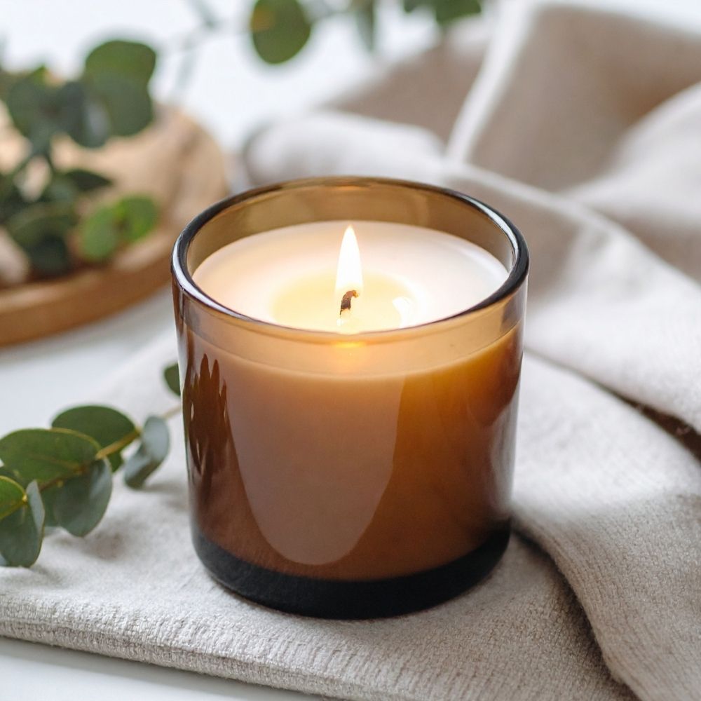 A lit amber glass candle jar with white wax sits on a light beige textured cloth, with blurred green leaves and a neutral background.