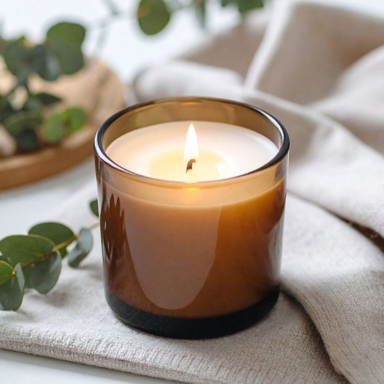 A lit amber glass candle jar with white wax sits on a light beige textured cloth, with blurred green leaves and a neutral background.