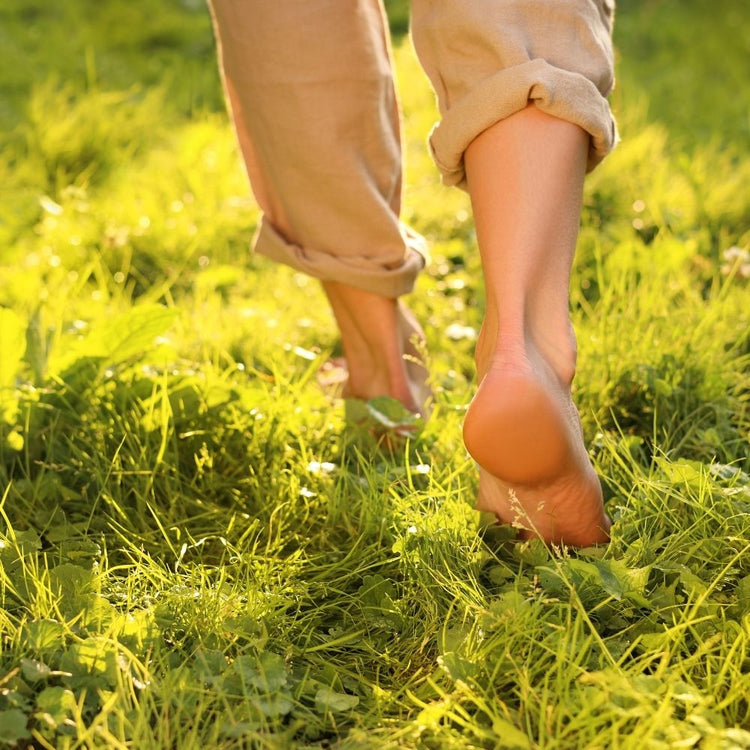 A close-up lifestyle shot of a person walking barefoot on bright green grass, wearing rolled-up beige pants.