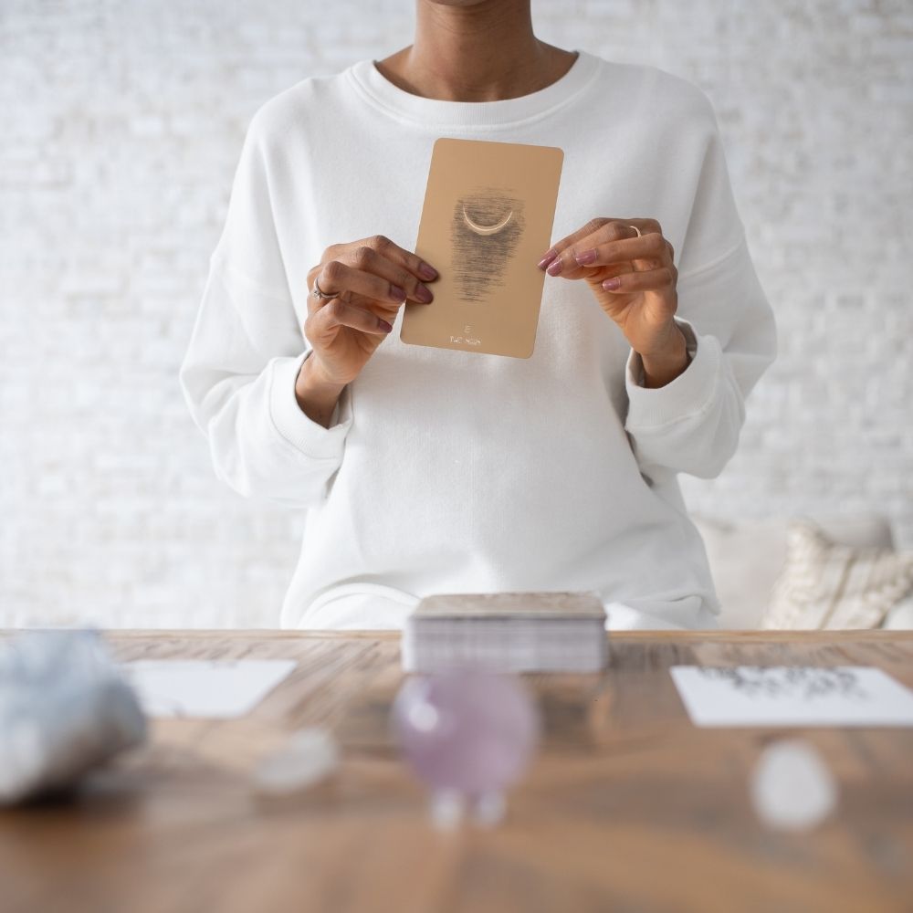 A person in a white sweatshirt holds up a tan oracle card with a crescent moon illustration, standing behind a wooden table with crystals and cards in a bright, modern room.