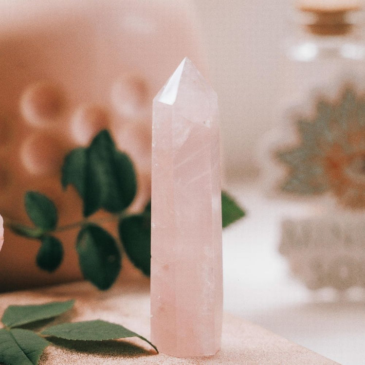 A pale pink crystal point tower stands upright on a soft beige surface, with blurred green leaves and a warm-toned decorative background.