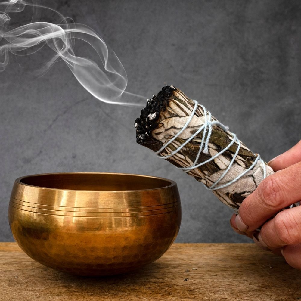 A brass singing bowl sits on a wooden surface with a hand holding a smudge bundle near it, and smoke curls against a dark gray background.