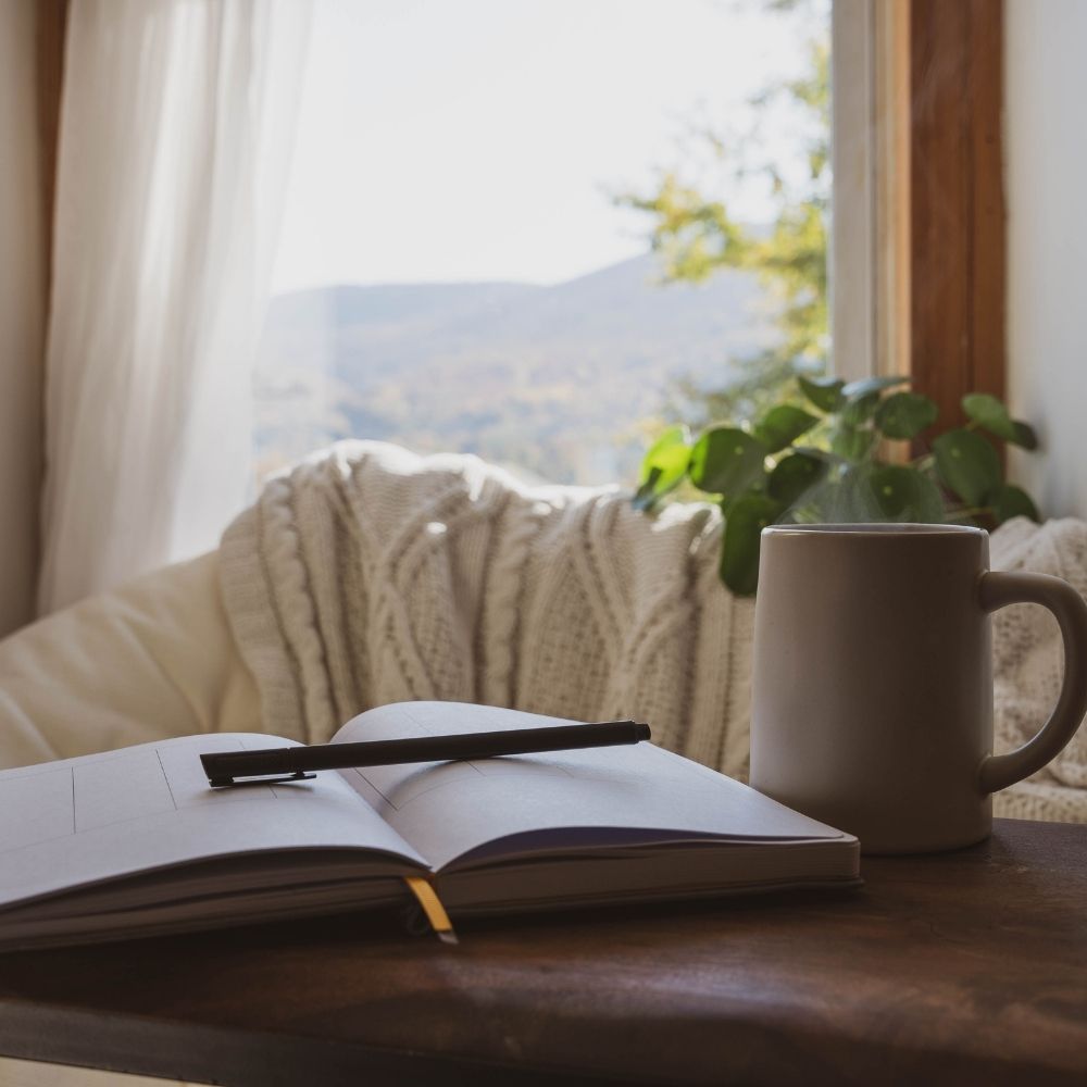 An open notebook with a black pen resting on it sits on a wooden table next to a beige ceramic mug, with a cozy cream cable-knit throw blanket and a potted plant in the foreground. A bright window behind shows a blurred mountain landscape.