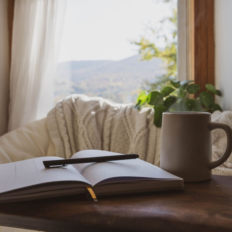 An open notebook with a black pen resting on it sits on a wooden table next to a beige ceramic mug, with a cozy cream cable-knit throw blanket and a potted plant in the foreground. A bright window behind shows a blurred mountain landscape.