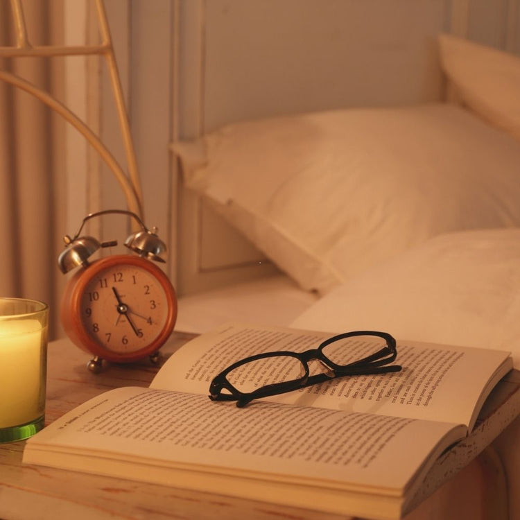 A pair of black-framed eyeglasses resting on an open book on a bedside table, with a lit candle, a twin-bell alarm clock, and a softly blurred bed in the background.