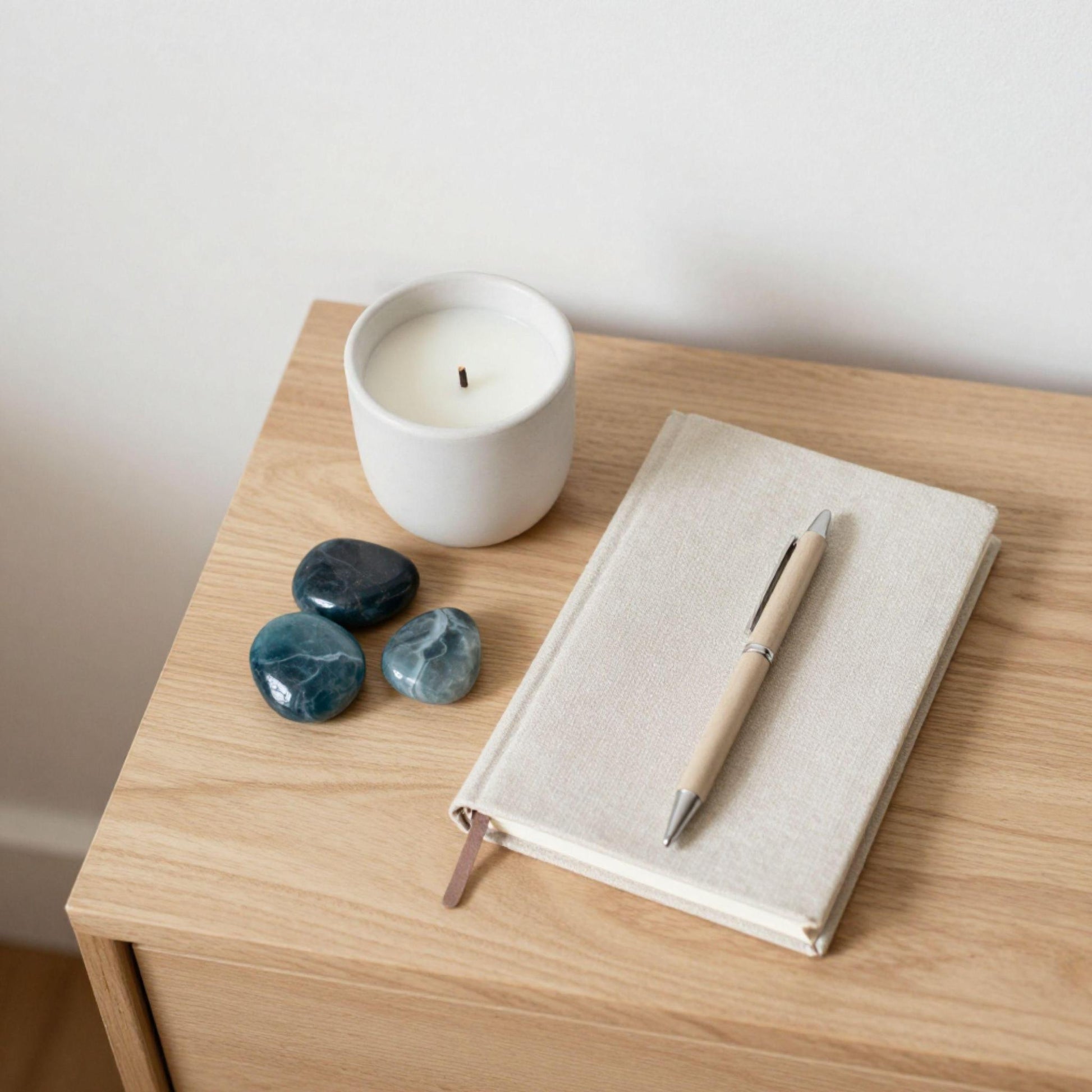 A beige canvas notebook with a pen resting on top, placed on a light wood nightstand next to a white candle, three blue-apatite polished stones, and a white wall.