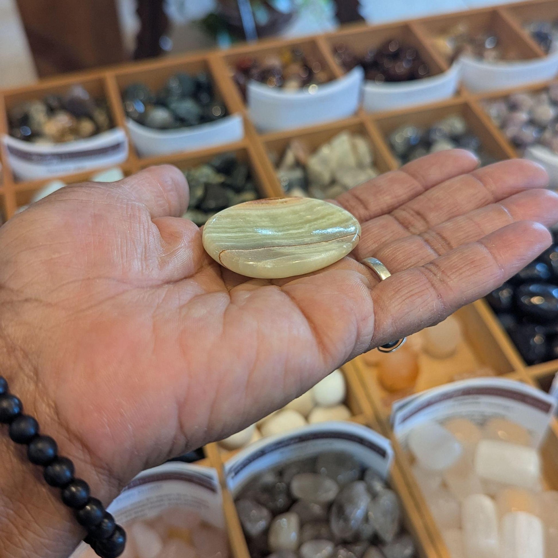 A polished oval, green onyx stone with layered beige, cream, and light brown bands rests on an open hand, with a blurred background of a crystal and gemstone display.