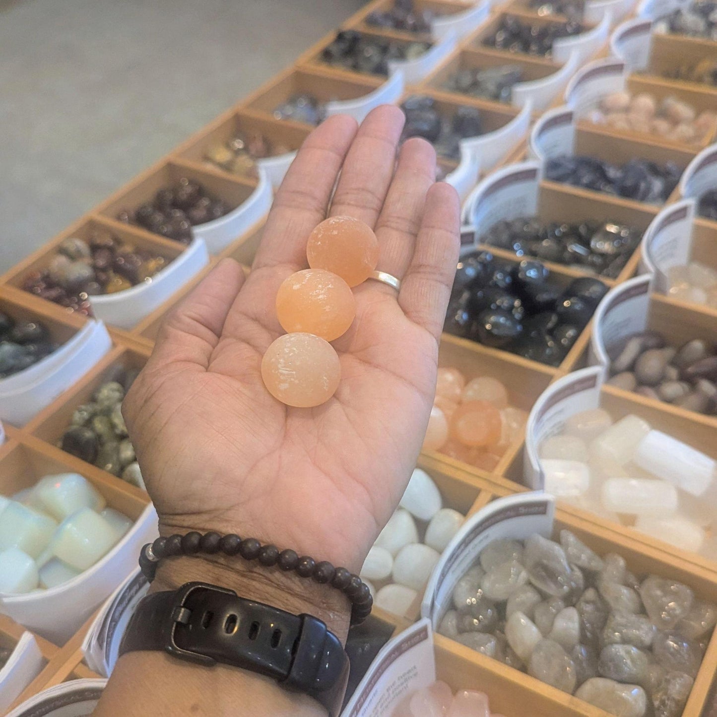 A hand holds three smooth, peach-colored crystal balls over a display of various gemstones and crystals in a shop.