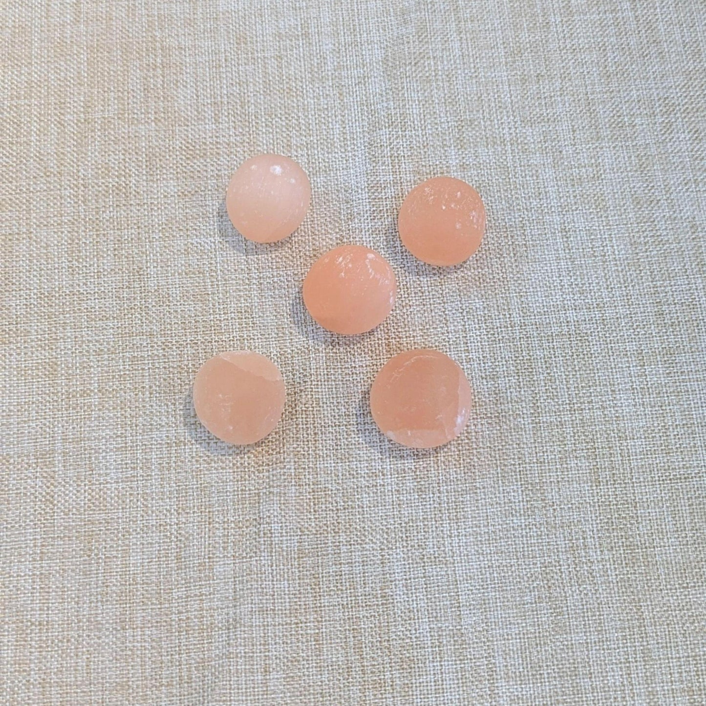 Five small round peach-colored (orange selenite) tumbled stones arranged on a textured beige fabric background.